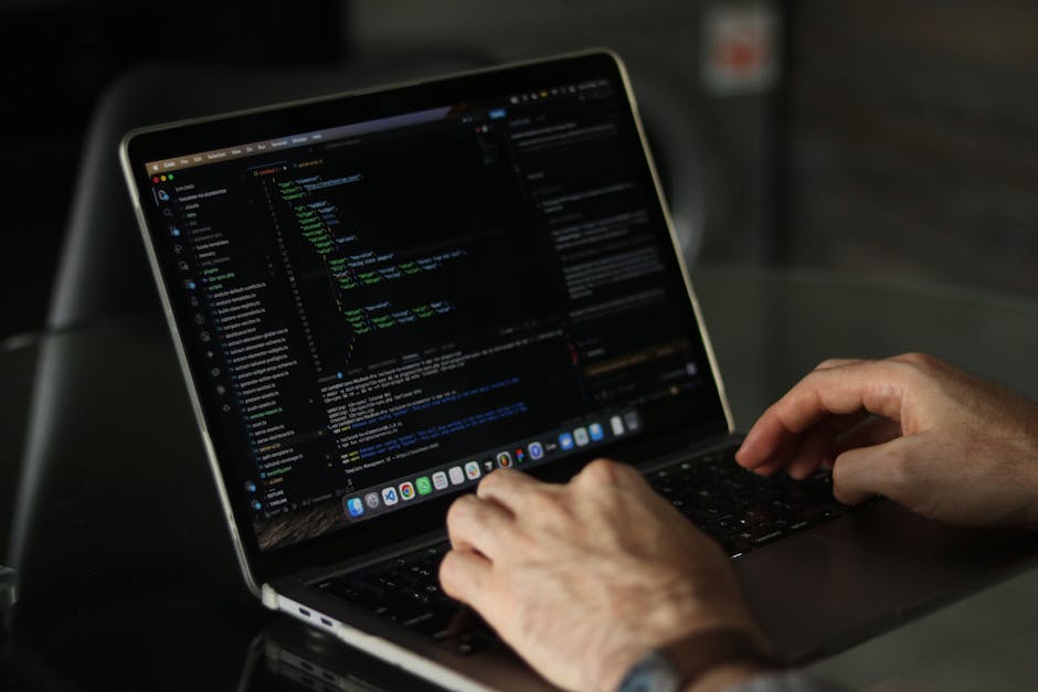 A person typing on a laptop with a dark screen displaying lines of colourful code, situated on a black table. The individual's hands are visible, with one hand resting on the keyboard and the other po
